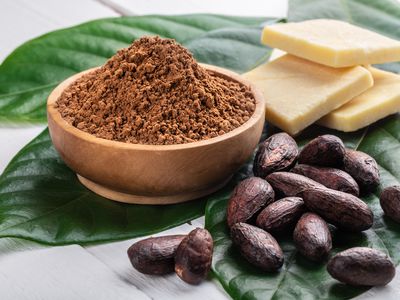 Cacao powder in wooden bowl, whole cacao beans, pieces of cocoa butter on white rustic table