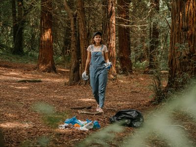 A woman in overalls picks up garbage in the forest.