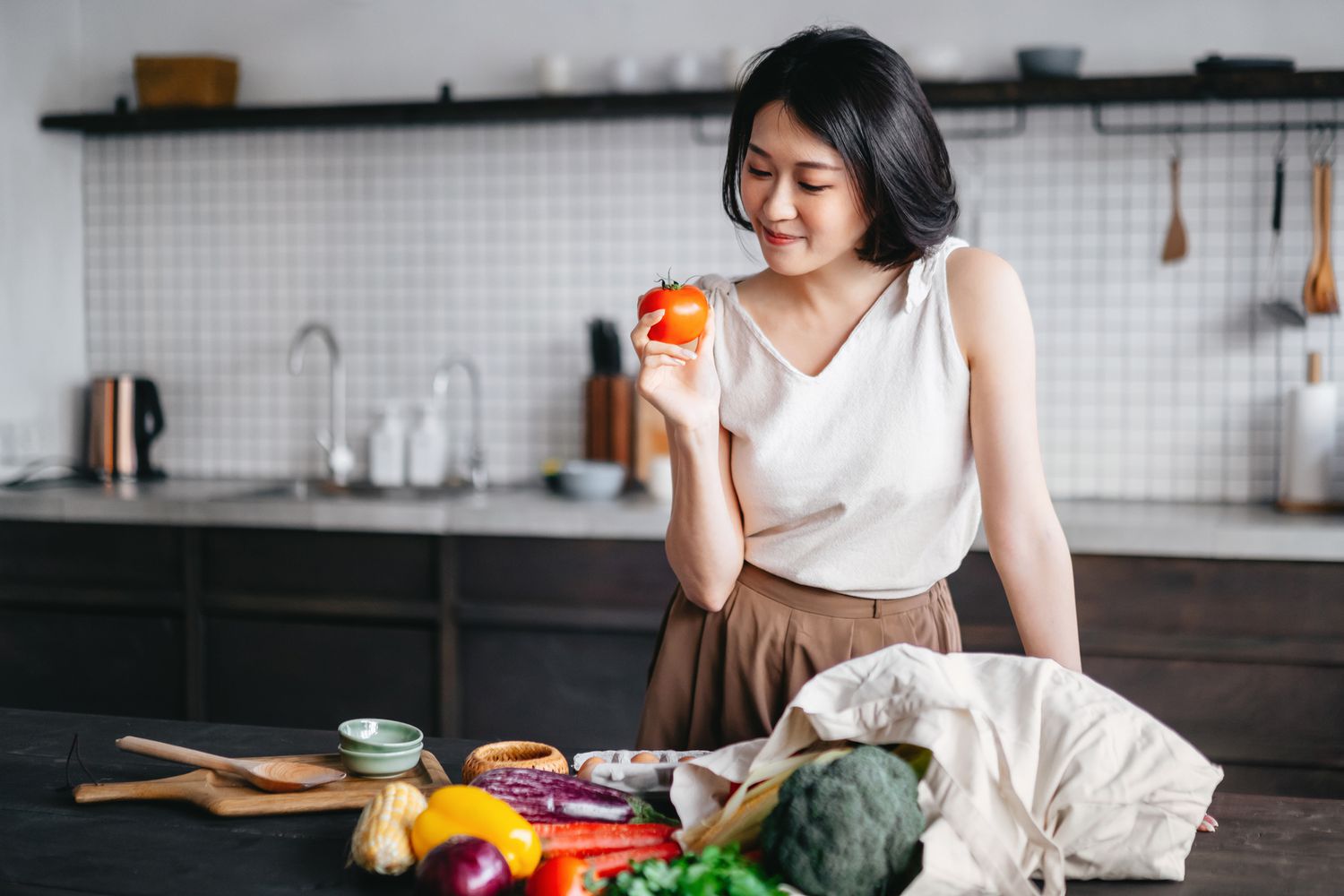 Young Asian woman coming home from grocery shopping and taking out fresh fruits and vegetables from a reusable shopping bag on the kitchen counter. She is planning to prepare a healthy meal with fresh produces