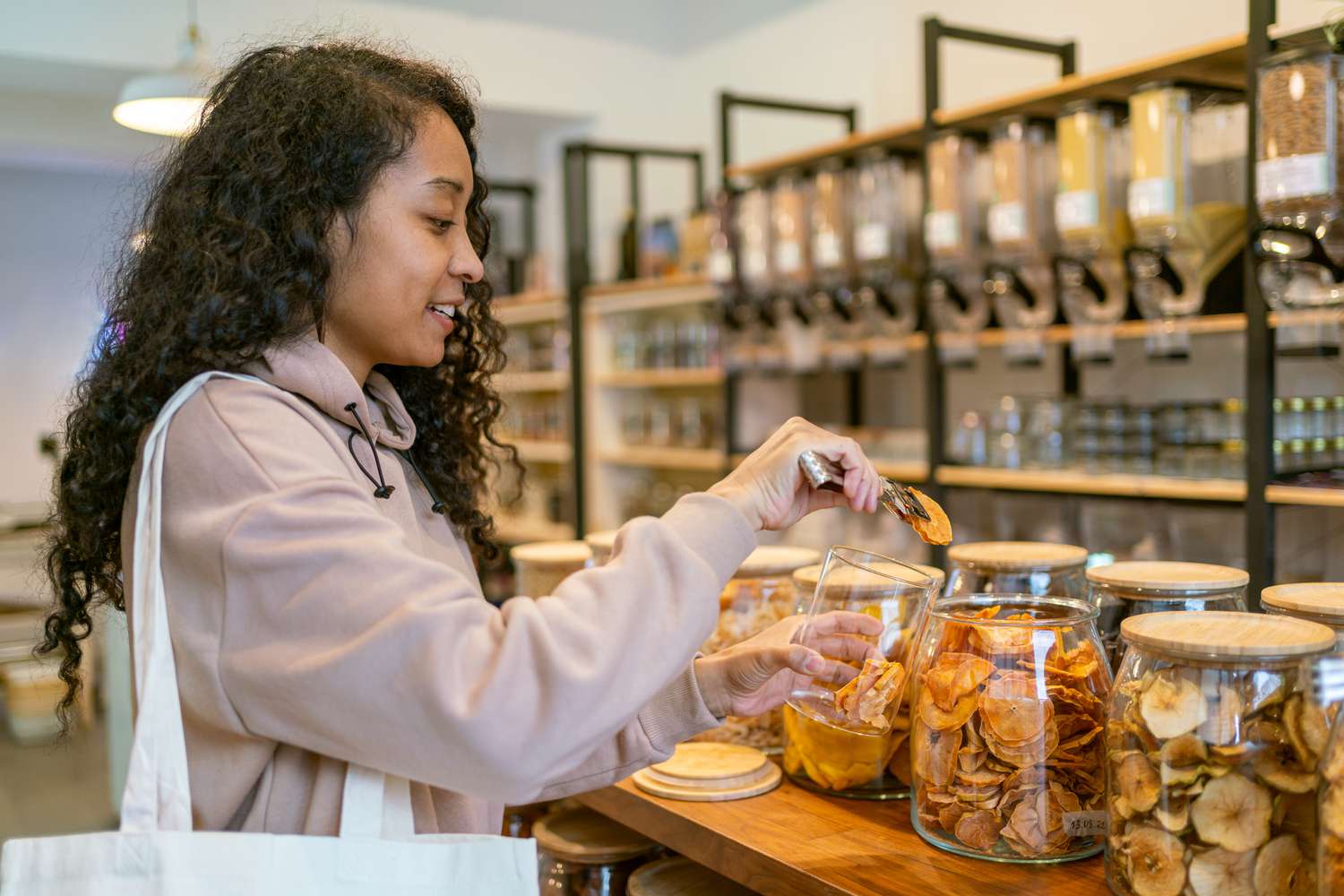 Young african-american female grabbing trail mix of citrus with tongs from glass jar in eco store