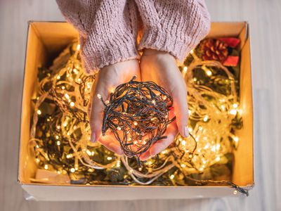 woman in pink sweater holds out bundle of glowing Christmas lights above full cardboard box