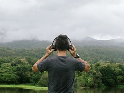 Rear View Of Man Listening Music While Looking At Mountains By Lake